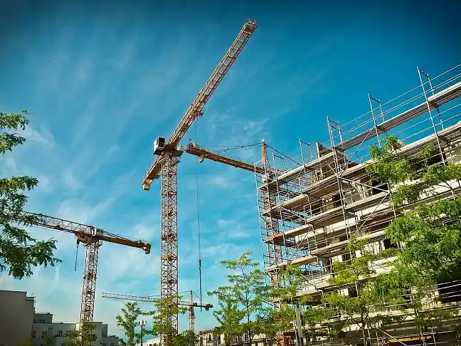 Abbildung einer Gebäude-Baustelle mit Kränen, im Vordergrund grüne Bäume.