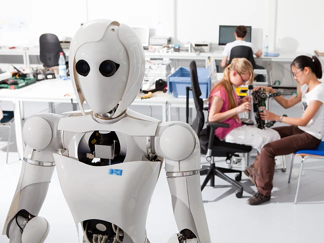 Weißer humanoider Roboter und im Hintergrund arbeiten Schülerinnen an einem technischen Modell