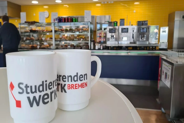 Zwei Tassen mit dem Aufschrift Studierendenwerk Bremen stehen auf einem Tisch in der Cafeteria